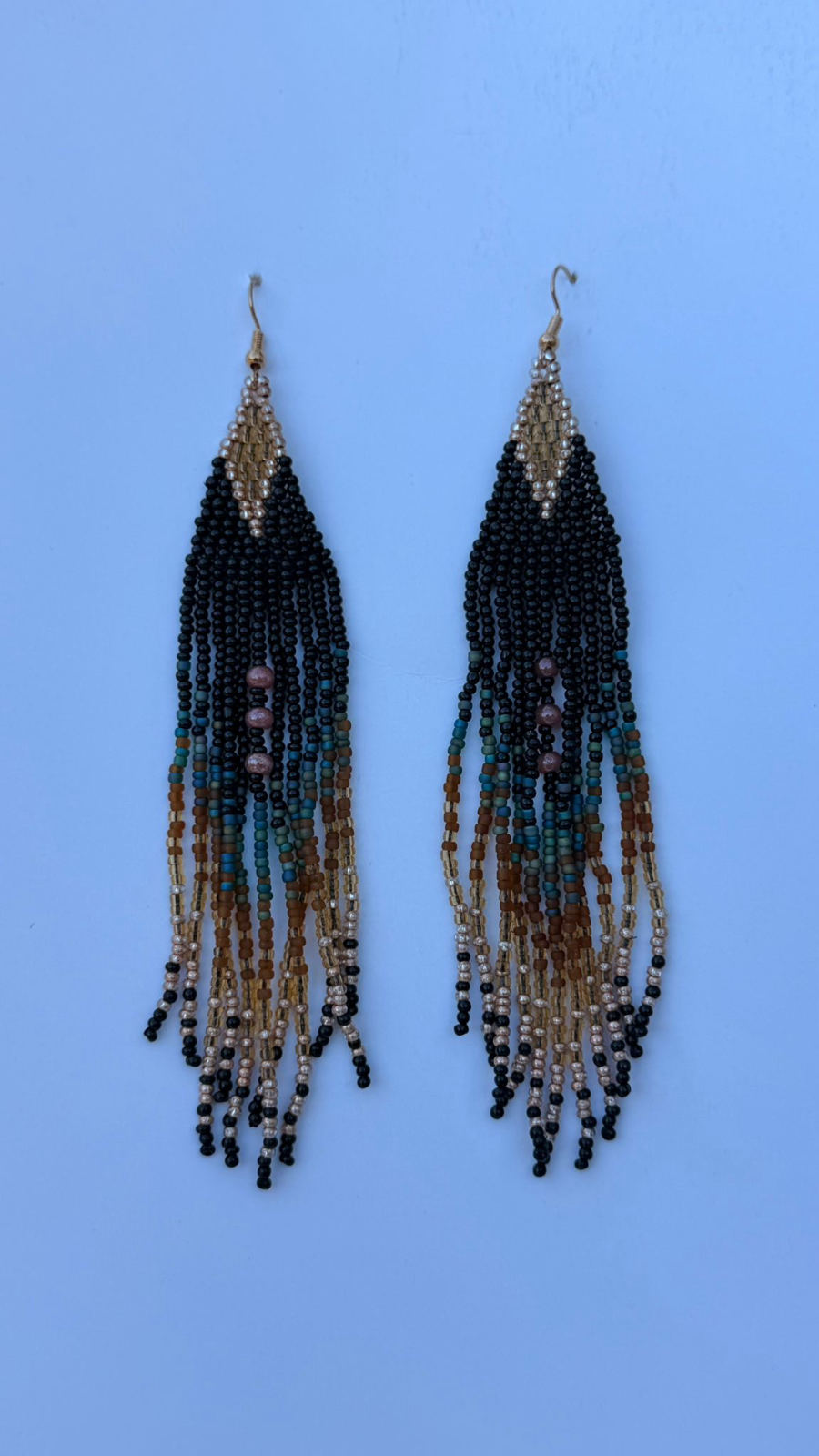 Long triangular fringe drop earrings with jet black triangle apex, fringe transitioning through dusty teal, warm caramel, champagne gold, and black seed bead tips with mauve round accent beads mid-fringe, gold fish-hook closures.