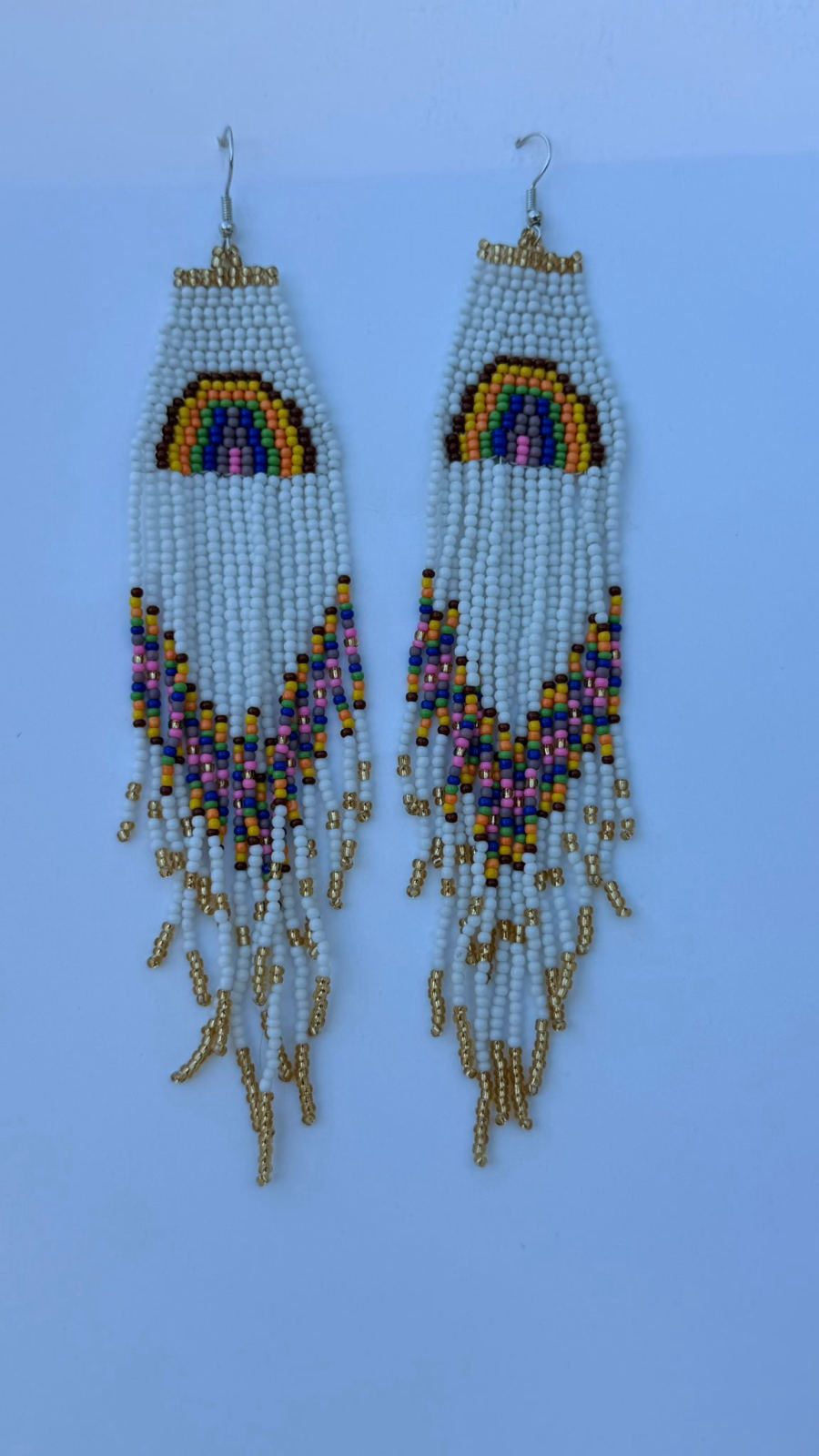 Long rectangular drop earrings with white seed bead panel featuring hand-stitched rainbow arc motif in yellow, brown, green, violet, blue, and pink, below which flows multicolor fringe in the same palette with gold metallic accents, silver fish-hook closures.
