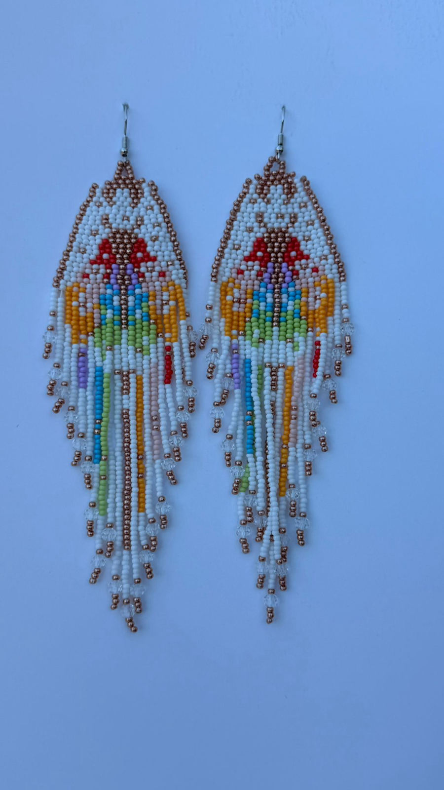 Long triangular fringe earrings with white seed bead panel featuring a figural motif in cherry red, marigold, sky blue, mint, and lavender, rose gold metallic border, crystal-dotted fringe strands in white and pastel colours, silver fish-hook closures.