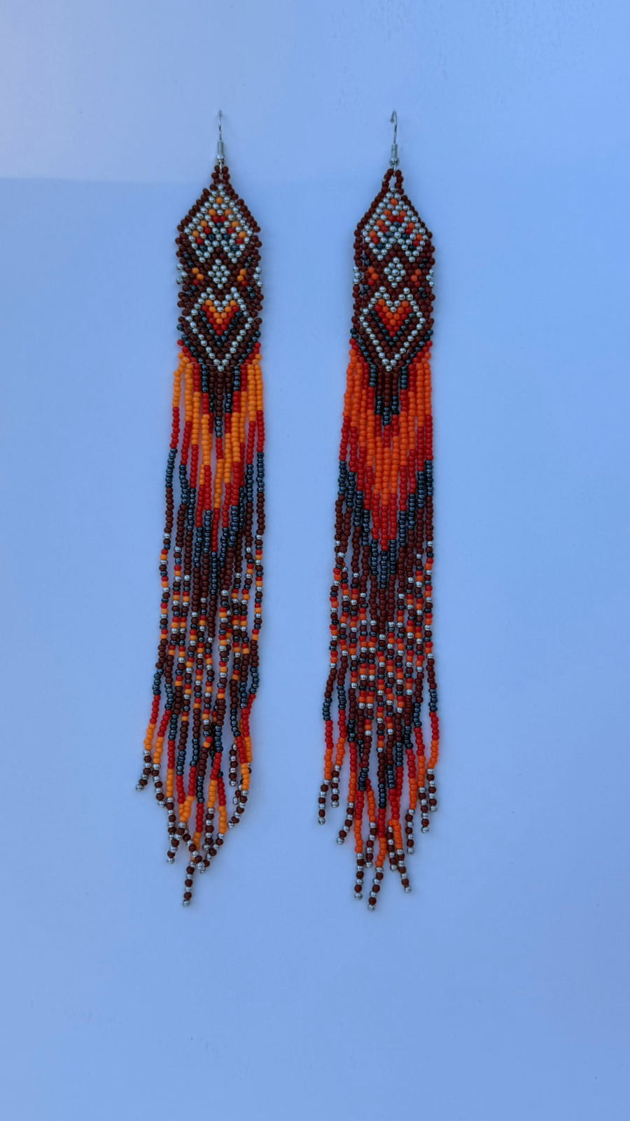 Very long fringe drop earrings with geometric diamond chevron header panel in oxblood, burnt orange, tangerine, pale silver-grey, and white seed beads, extra-long fringe repeating the same chevron color pattern with silver metallic accent beads at the tips, silver fish-hook closures.