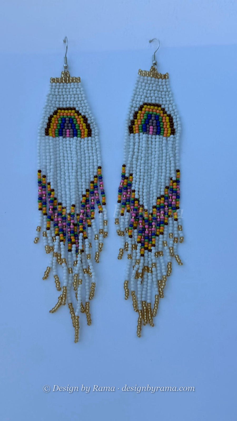 Long rectangular drop earrings with white seed bead panel featuring hand-stitched rainbow arc motif in yellow, brown, green, violet, blue, and pink, below which flows multicolor fringe in the same palette with gold metallic accents, silver fish-hook closures.